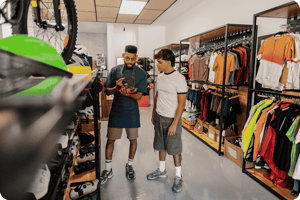A store employee wearing an apron shows a red and black cycling shoe to a customer inside a bicycle apparel shop.